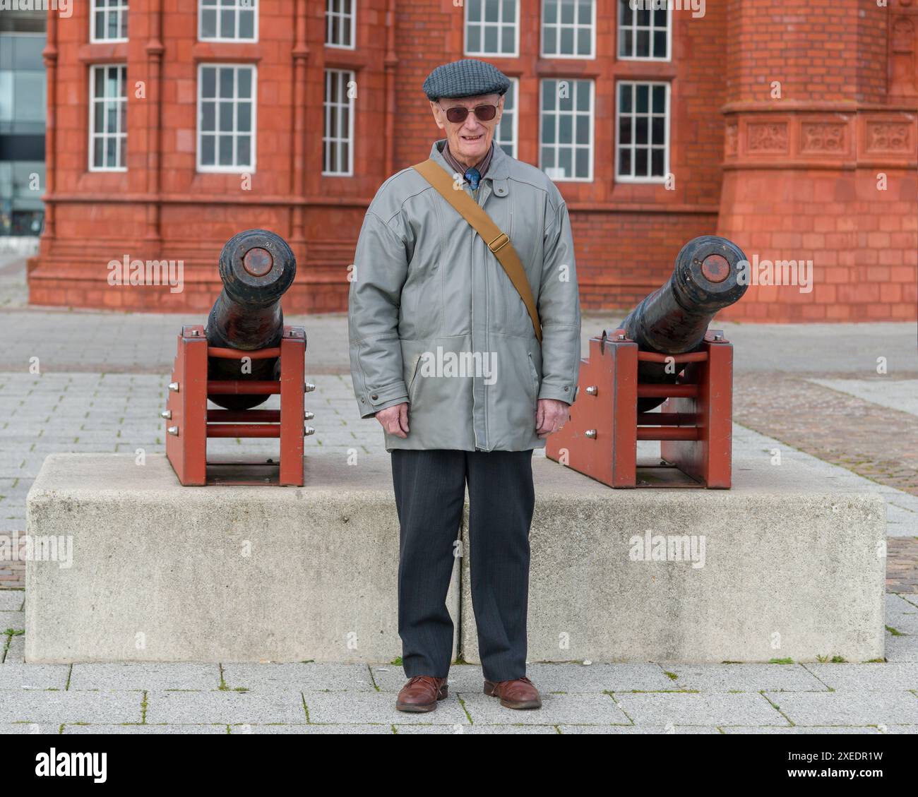 Ein alter Soldat in grauer Lederjacke, dunkler Hose, Flatcap und Sonnenbrille erinnert sich an seinen nationalen Dienst und steht zwischen zwei Kanonen. Stockfoto