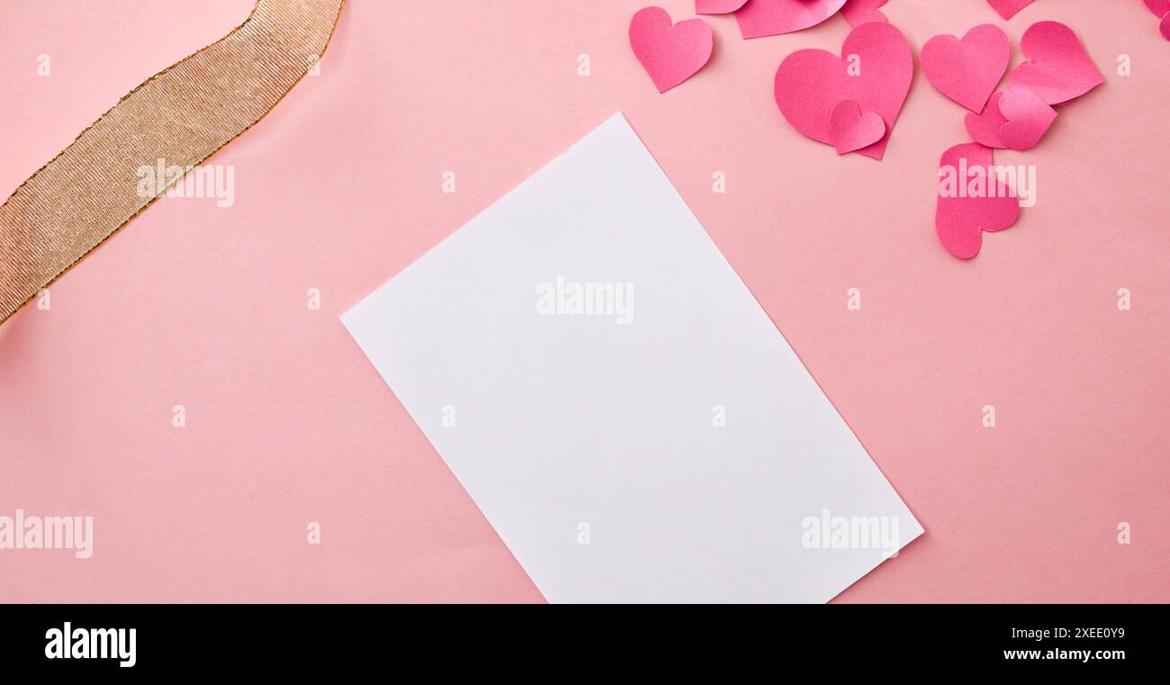 Hintergrund mit leerer Karte auf rosafarbenem Tisch mit herzförmigen Papierausschnitten und goldenem dekorativem Stoffband. Draufsicht. Stockfoto