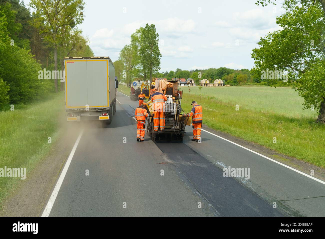 Tarnow, Polen - 19. Mai 2023: Straßenarbeiter in reflektierenden Westen bedienen auf einer Landstraße Straßenpflastermaschinen mit vorbeifahrenden Fahrzeugen. Stockfoto