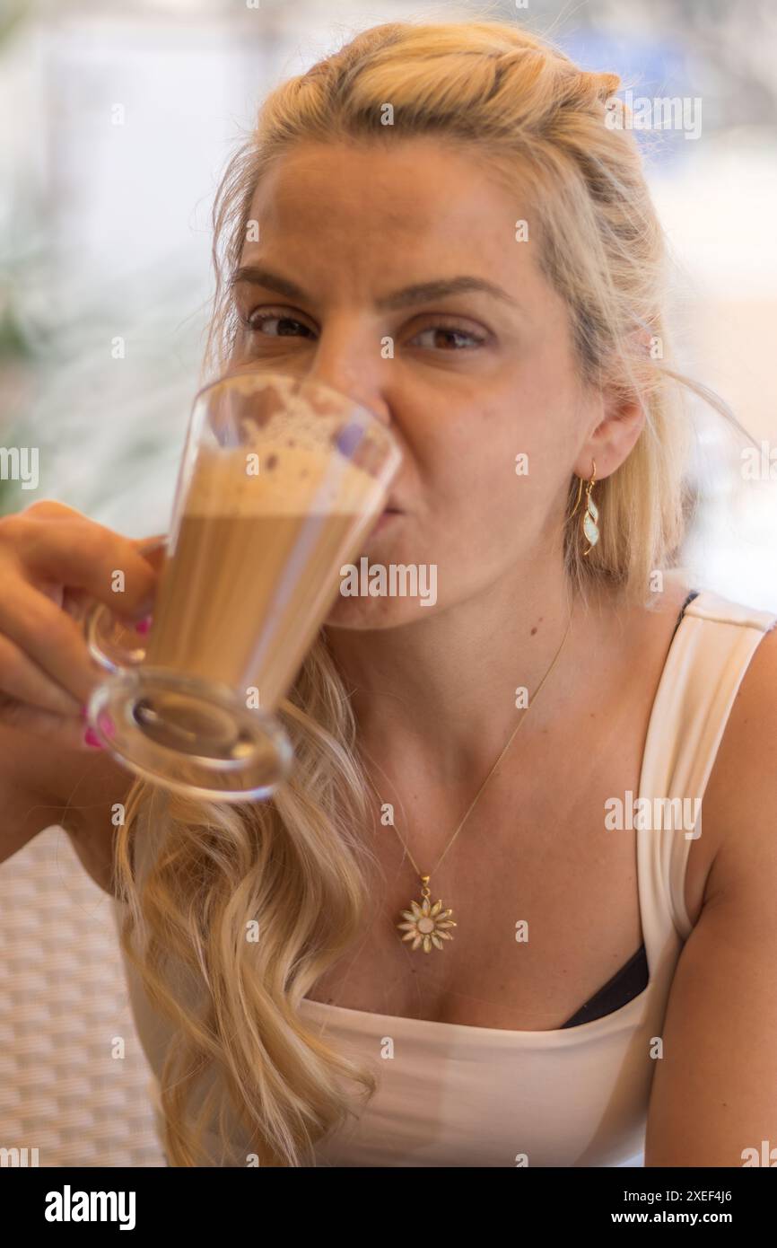 Dieses Bild zeigt eine schöne blonde Frau, die einen Cappuccino genießt. Sie scheint an einem Tisch in einer ungezwungenen, sozialen Umgebung zu sitzen Stockfoto