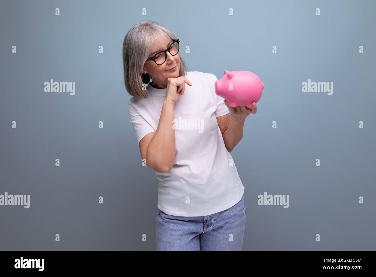 Eine Frau im Alter mit grauen Haaren hält Geldkapital von einer Schweinebank auf einem Atelierhintergrund mit Kopierraum Stockfoto