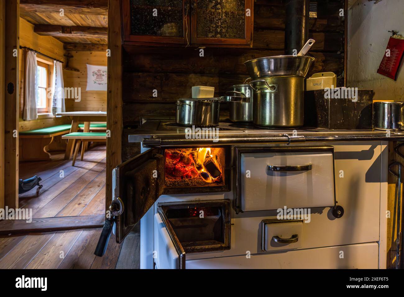 Ein alter Holzofen steht im sogenannten ‚Vorkaser‘, dem traditionellen Wohn- und Arbeitsraum einer Almhütte, auf der Filzmoosalm bei Großarl, Salzburg, Österreich. Mehrere Kochtöpfe warten auf der Kochplatte. Der holzvertäfelte Wohnraum mit einem Tisch und Bänken ist im Hintergrund zu sehen. Stockfoto