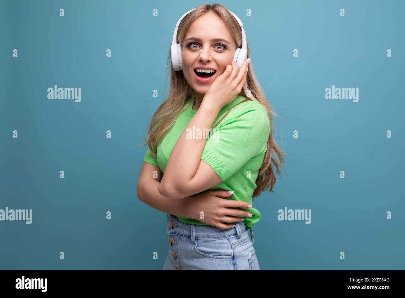 Das charmante blonde Mädchen im lässigen Outfit hört Musik in großen weißen Kopfhörern auf blauem Hintergrund Stockfoto