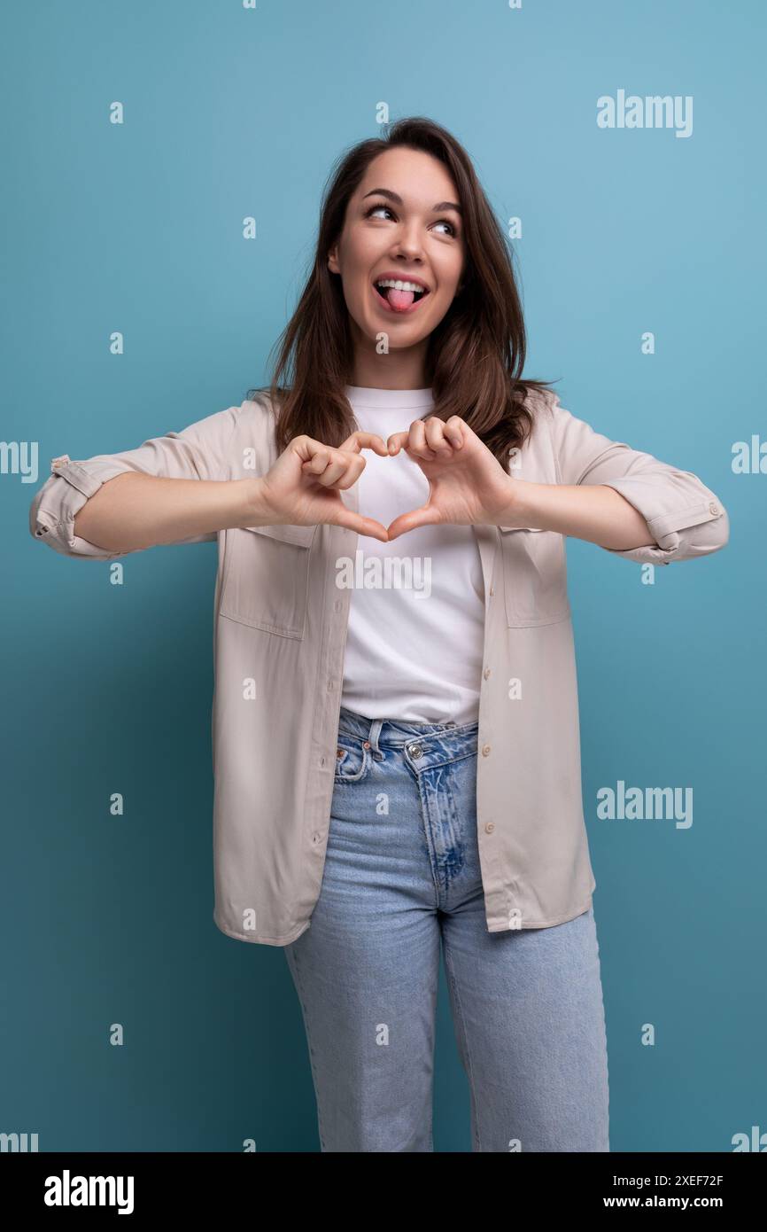 30s Frau mit schick schwarzem Haar unter den Schultern in einem beigen Hemd zeigt ein Herz mit ihren Händen Stockfoto