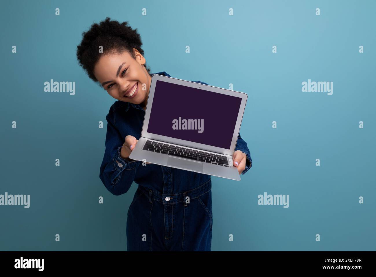Junge fröhliche hispanische Geschäftsfrau mit flauschigem Haar in blauem Denim-Anzug zeigt Laptop-Bildschirm mit Mockup Stockfoto