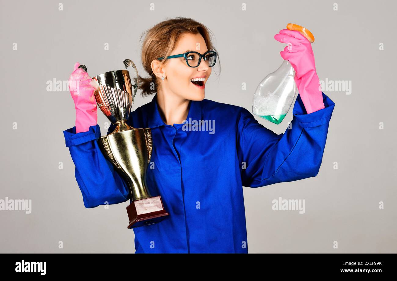 Professioneller Reinigungsservice. Lächelnde Frau in Uniform und Gummihandschuhen mit goldenem Trophäenbecher und Reinigungsspray. Reinigung, Sauberkeit, Reinigungsprofi Stockfoto