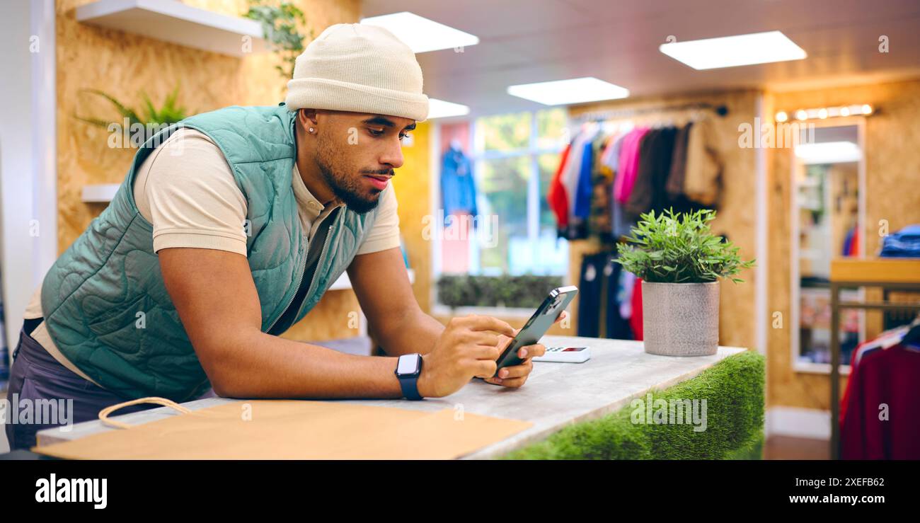 Männlicher Vertriebsmitarbeiter Am Kassenschalter Im Pop-Up-Mode- Oder Kleiderladen, Der Sich Das Mobiltelefon Anschaut Stockfoto