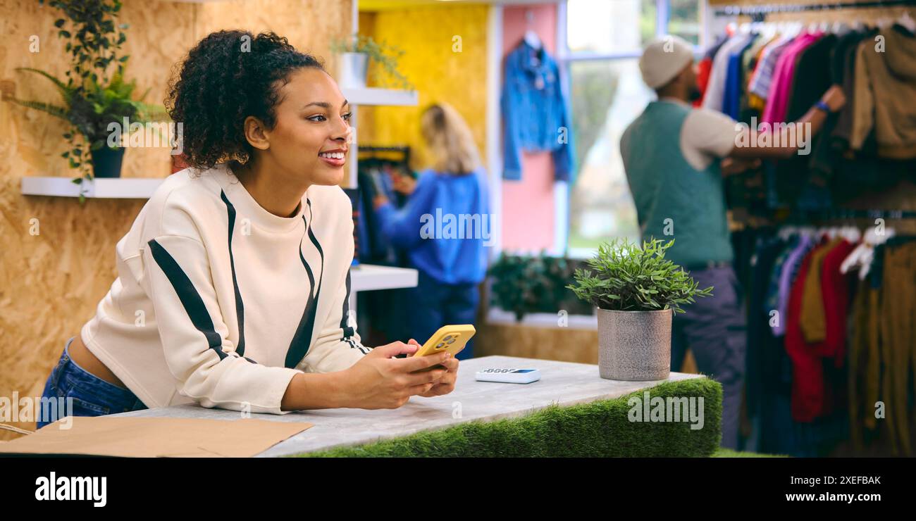 Weibliche Verkäuferin An Der Kasse Im Pop-Up-Mode- Oder Kleidergeschäft, Die Sich Ein Mobiltelefon Anschaut Stockfoto