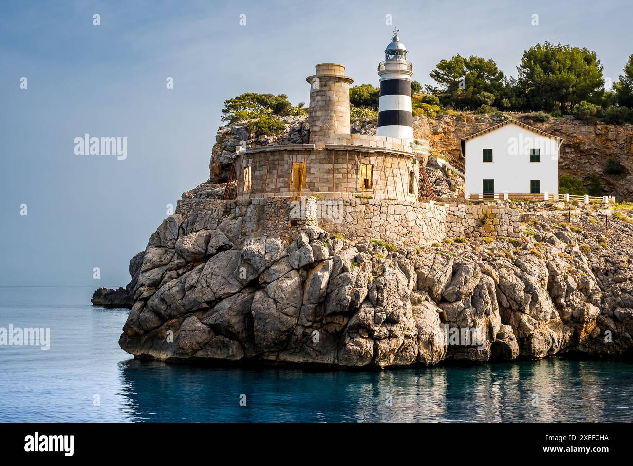 Der Leuchtturm von SA Creu, der in der sanften Morgensonne getaucht ist, überblickt das ruhige Wasser von Port de Sóller, einer alten Leuchtturm-Ruine. Stockfoto