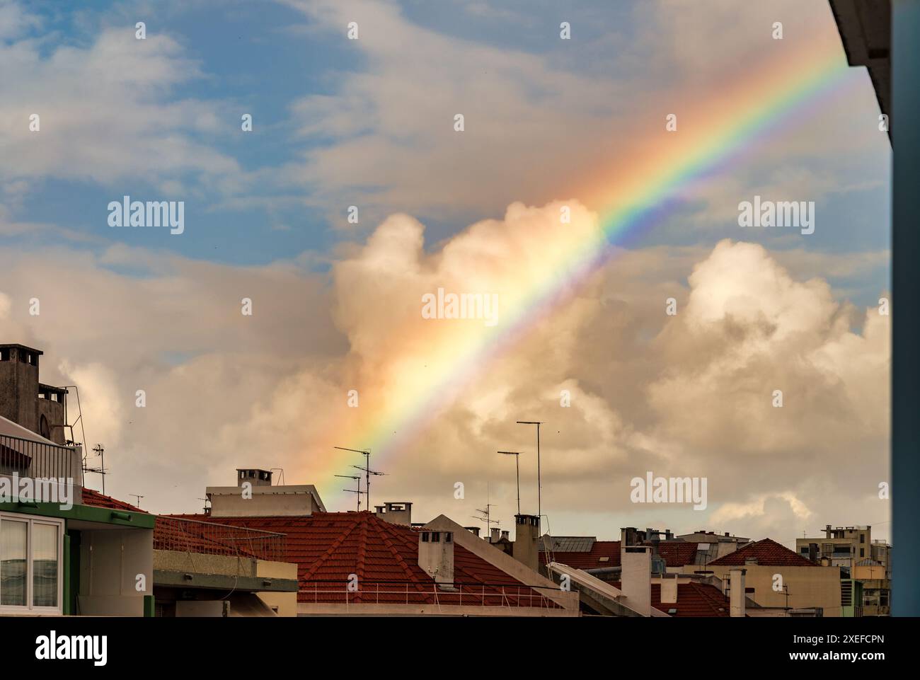 Ein lebhafter Regenbogenbogen zieht sich über die Dächer der Stadt und bildet einen Kontrast zu einem teilweise bewölkten Himmel und Stadtgebäuden. Stockfoto