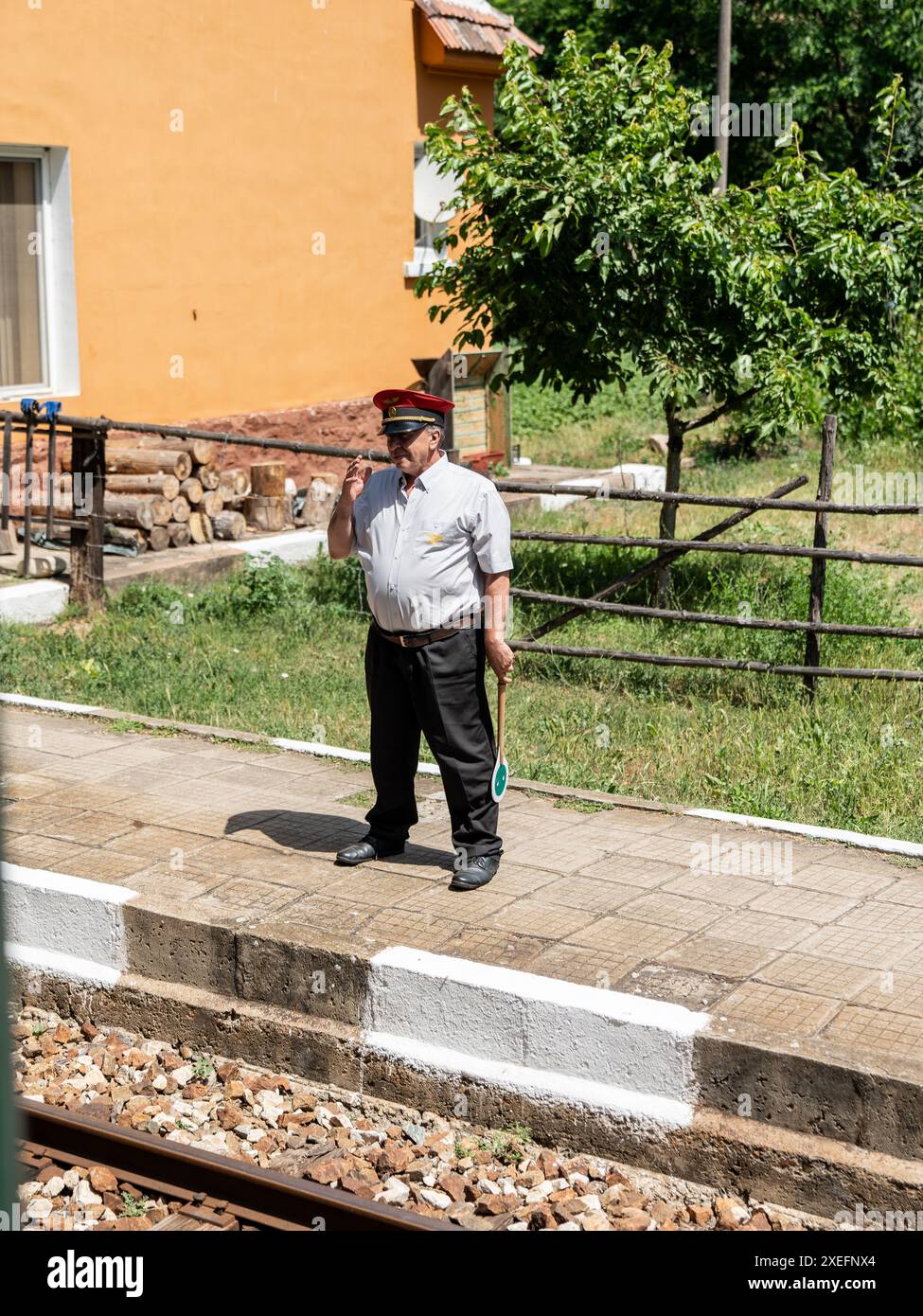 Bahnhofsmeister wartet auf die Ankunft des Zuges an einem kleinen Landbahnhof mit grüner Signalflagge an sonnigen Sommertagen Stockfoto