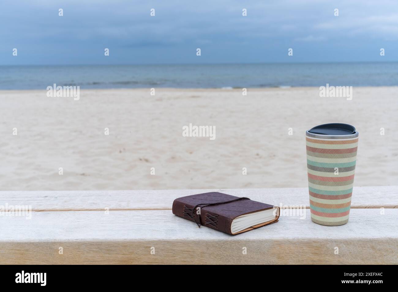 Eine Tasse Kaffee und ein Buch liegen auf einer Bank am Strand vor dem Hintergrund des Meeres. Postkartenurlaub auf dem Meer. Stockfoto