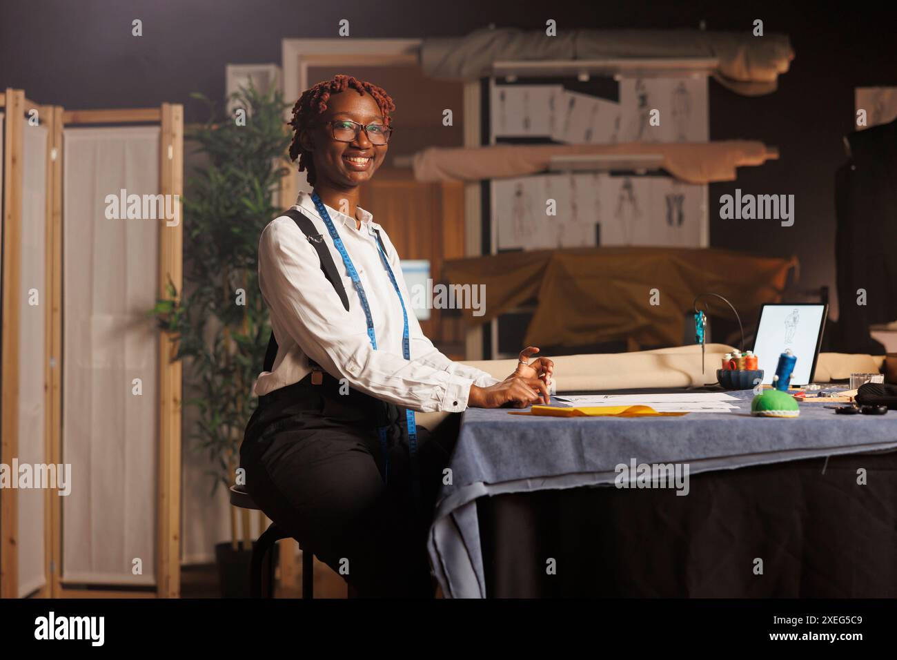 Porträt einer glücklichen afroamerikanischen Schneiderin, die in einem schwarzen Ateliergeschäft arbeitet. Professioneller, lächelnder Couturier, der maßgeschneiderte Outfits für die kommende Komission entwirft Stockfoto