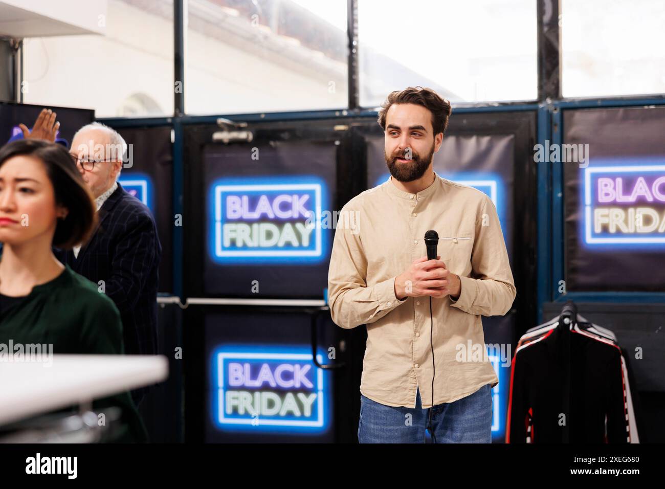 Junger Mann, lokaler TV-Reporter, der ein Mikrofon hält, das während des Black Friday-Verkaufs Live-Übertragungen vom Modegeschäft macht. Männlicher Korrespondent, der die Szene im Einkaufszentrum an dem geschäftigsten Einzelhandelstag des Jahres bedeckte Stockfoto