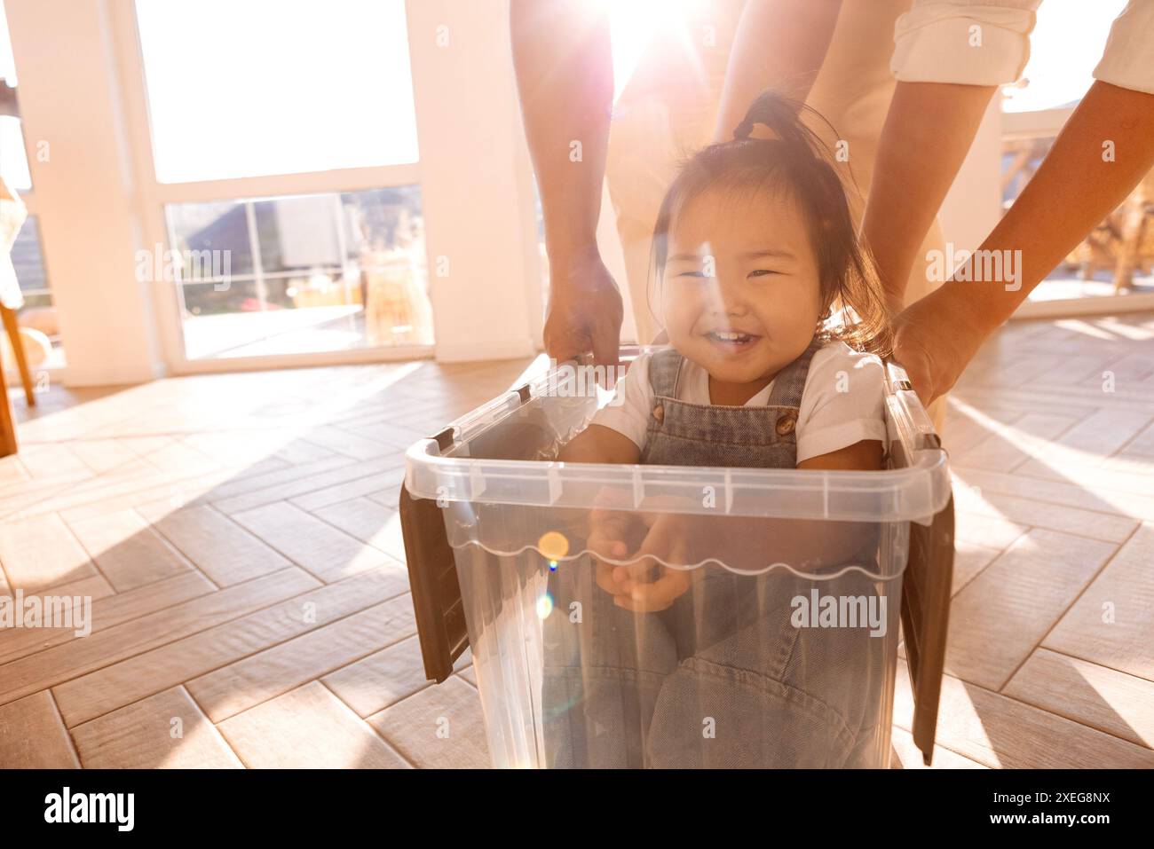 Junge lachende asiatische Eltern Rollen ihre Tochter in einer Plastikkiste um das Haus Stockfoto