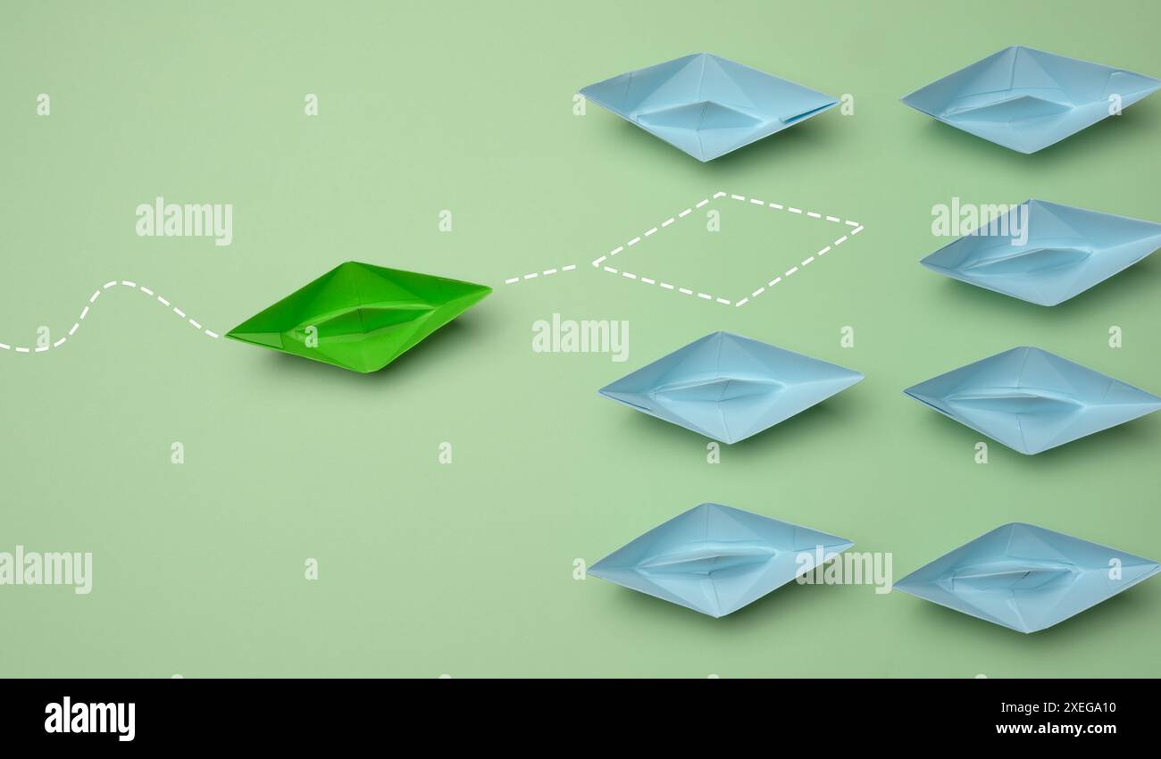 Gruppe blauer Papierboote, die in eine Richtung fahren, und eines grünes in die entgegengesetzte Richtung. Stockfoto