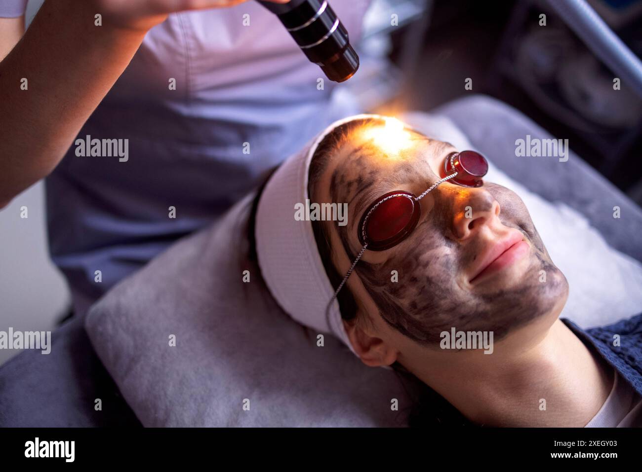 Kosmetikerin, die Schönheitslaser in der Nähe des Frauengesichts hält, bedeckt mit Kohlemaske Stockfoto