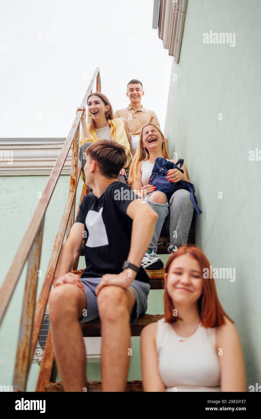 Lustige lächelnde Teenager in lässiger Kleidung sitzen auf einer Metallleiter auf dem Dach eines Gebäudes. Süße Freunde haben Spaß miteinander Stockfoto