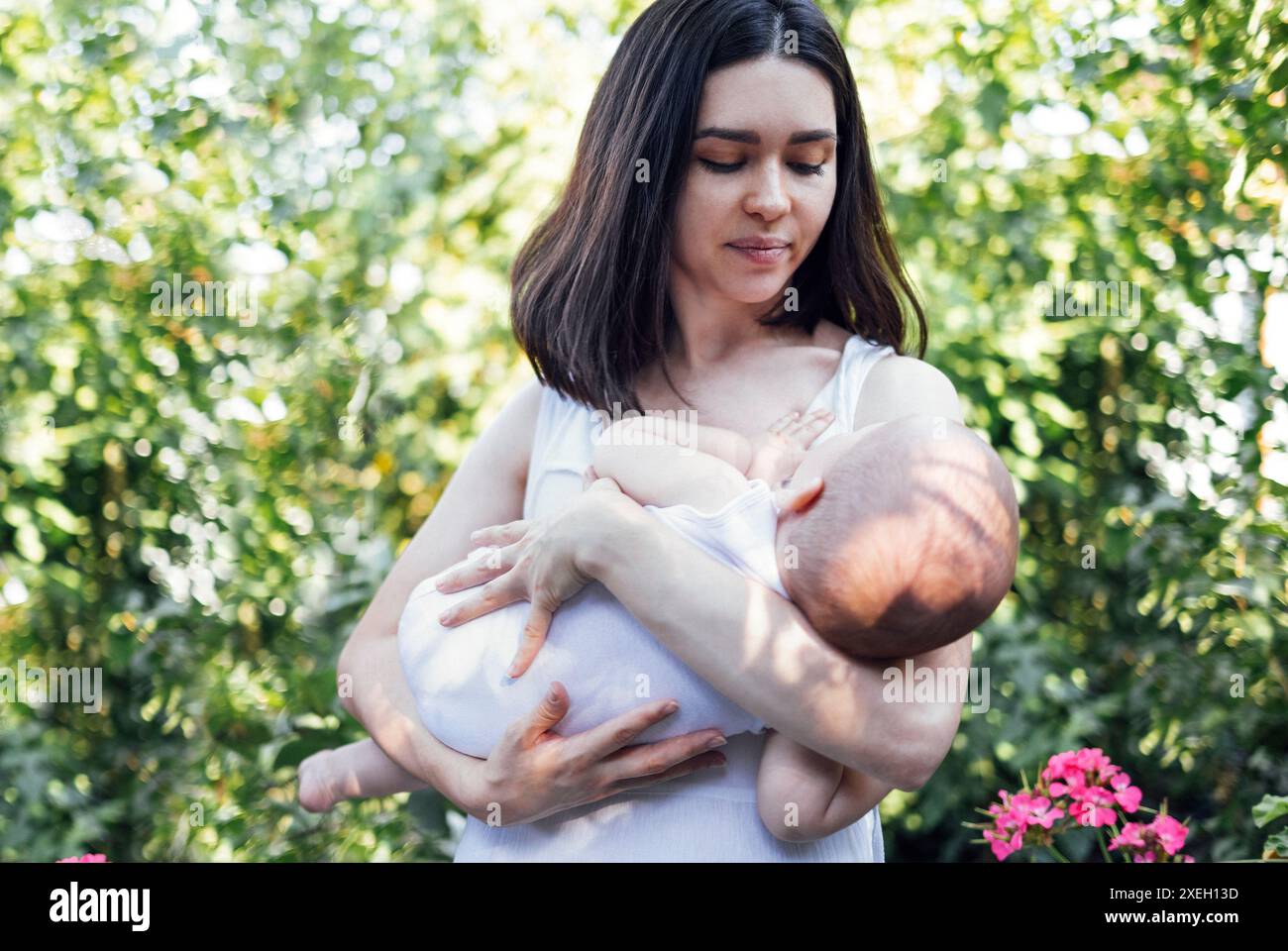 Eine schöne Mutter in weißen Kleidern schaukelt ihr Baby im Garten in den Armen. Stockfoto