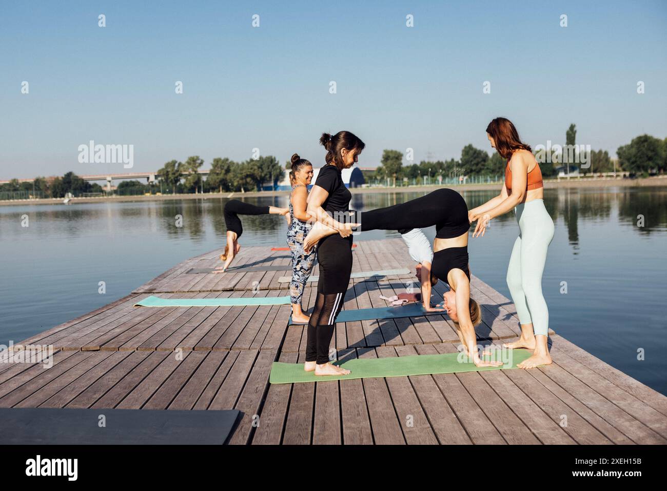Ein Yoga-Kurs zu zweit mit einem Lehrer auf einem hölzernen Pier am Fluss in der Stadt. Eine Gruppe von Frauen trainiert draußen am Morgen. Stockfoto