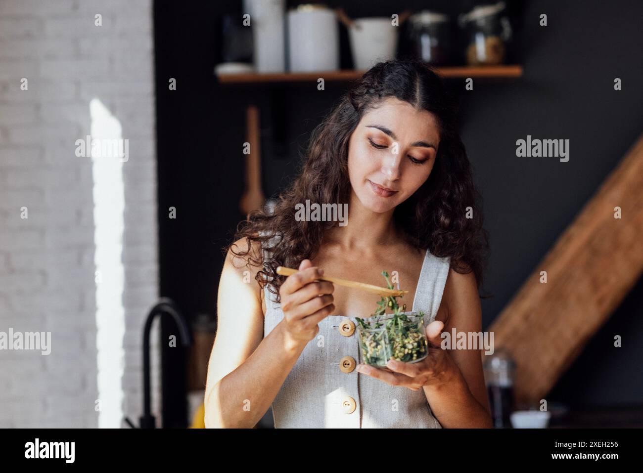 Attraktive junge Frau isst Salat aus gekeimtem Gemüse in der Küche. Lockiges, brünettes Mädchen in Leinenkleid hält Gabel an Stockfoto