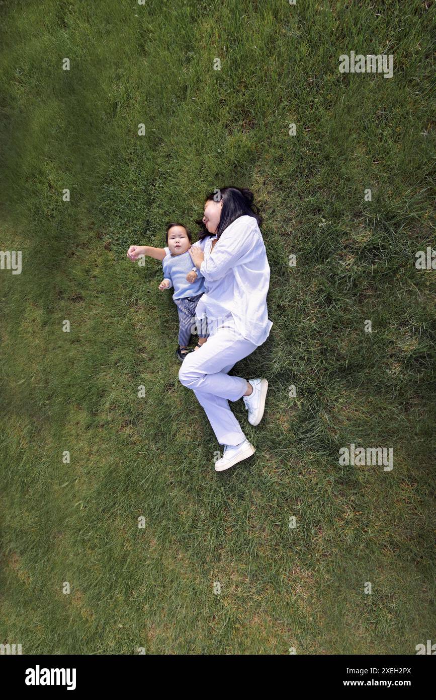 Asiatische lächelnde Mutter und süße Tochter liegen auf einem grünen Rasen. Eine charmante Koreanerin und ihr süßes Kind haben Spaß am G Stockfoto