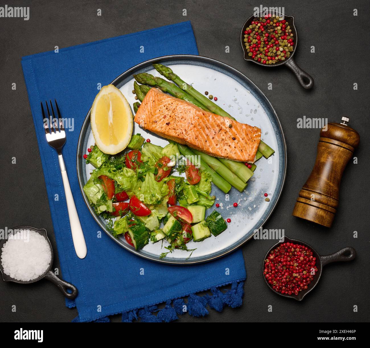 Gesundes Mittagessen mit gegrilltem Lachs mit Spargel und frischem Tomaten- und Gurkensalat, einer Zitronenscheibe. Essen auf einem blauen runden Teller Stockfoto