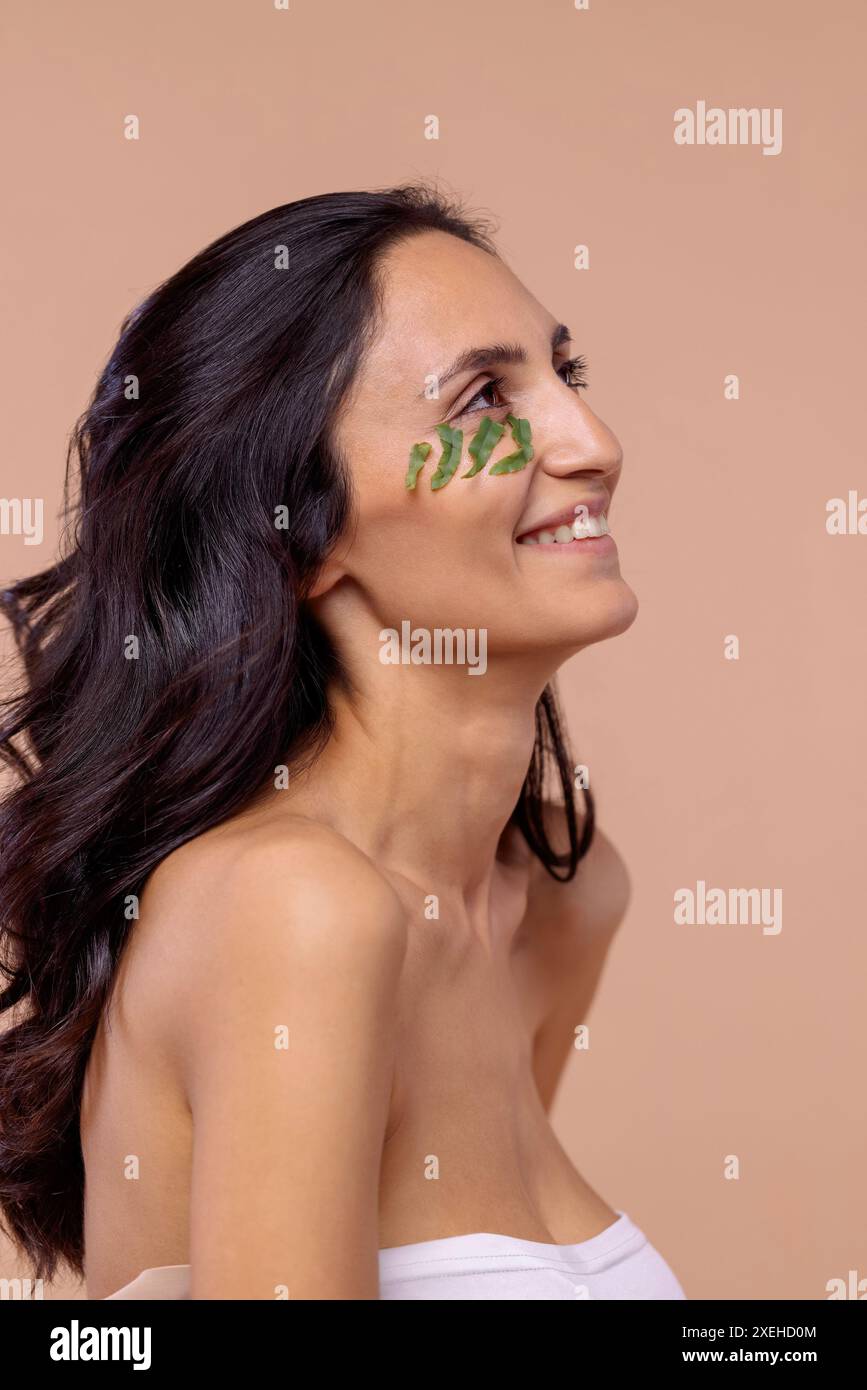 Studio-Porträt einer jungen Frau mit Blättern unter dem Auge auf einem beigefarbenen isolierten Hintergrund. Natürliche, umweltfreundliche Kosmetik. Takin Stockfoto