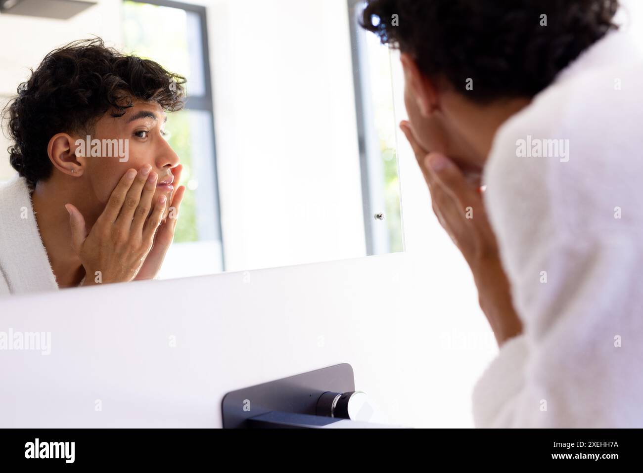 In den Spiegel schauen, junger Mann das Gesicht berühren und die Haut im Badezimmer untersuchen Stockfoto