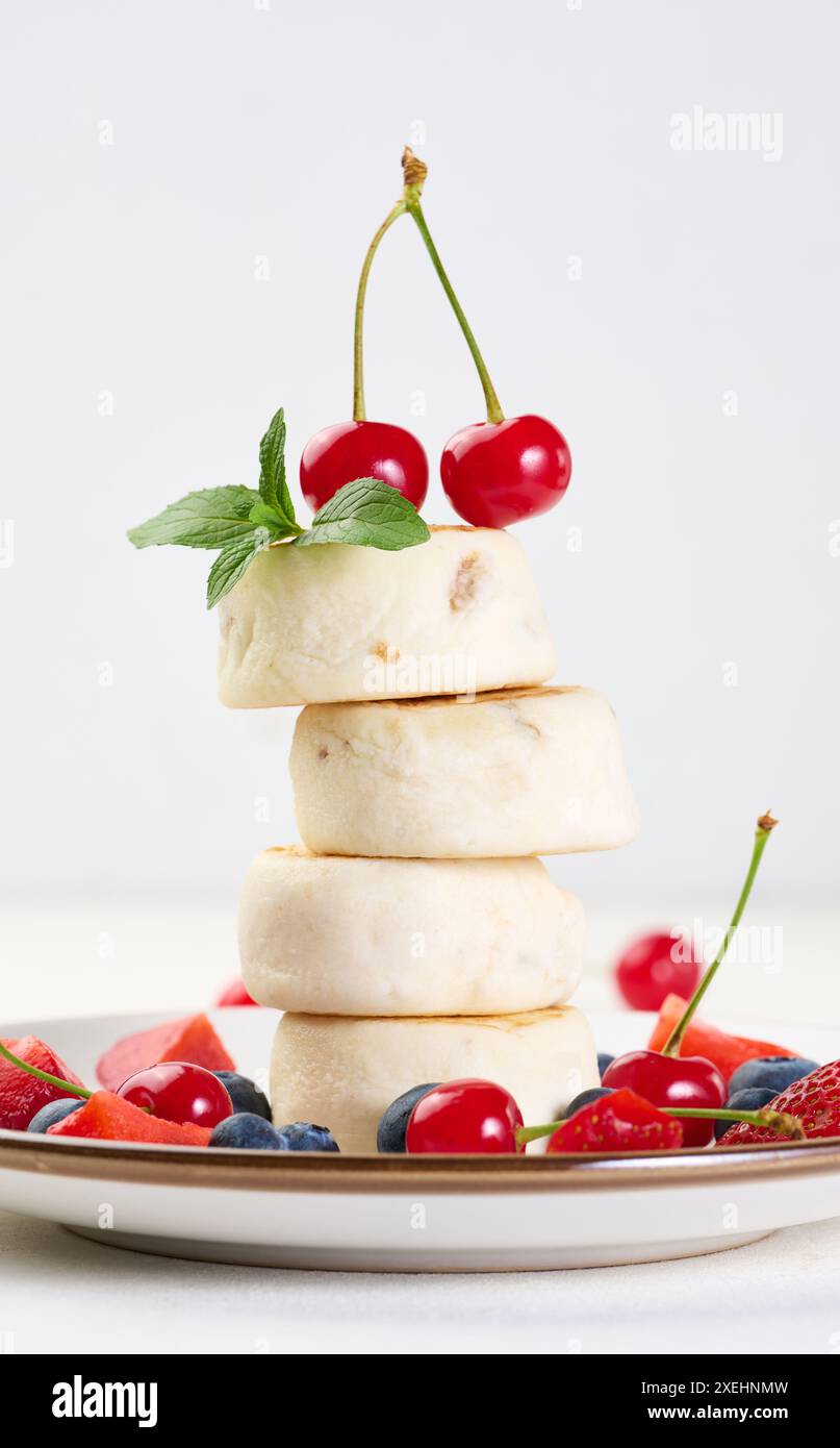 Ein Stapel von vier Käsekuchen, dekoriert mit zwei Kirschen und einem Minzzweig, auf einem Teller mit Beeren, Heidelbeeren, Erdbeeren Stockfoto