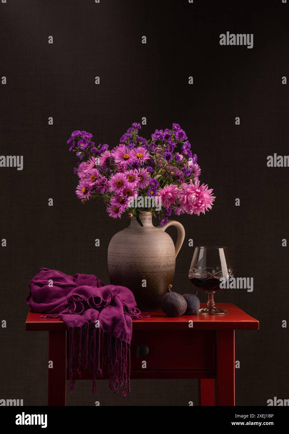 Blumenstrauß im Herbst. Viele lilafarbene Astern in einer braunen Vase. Neben einem Glas Rotwein auf einem roten Holztisch. Zwei frische Feigen. Dunkelrosa Schal. Stockfoto