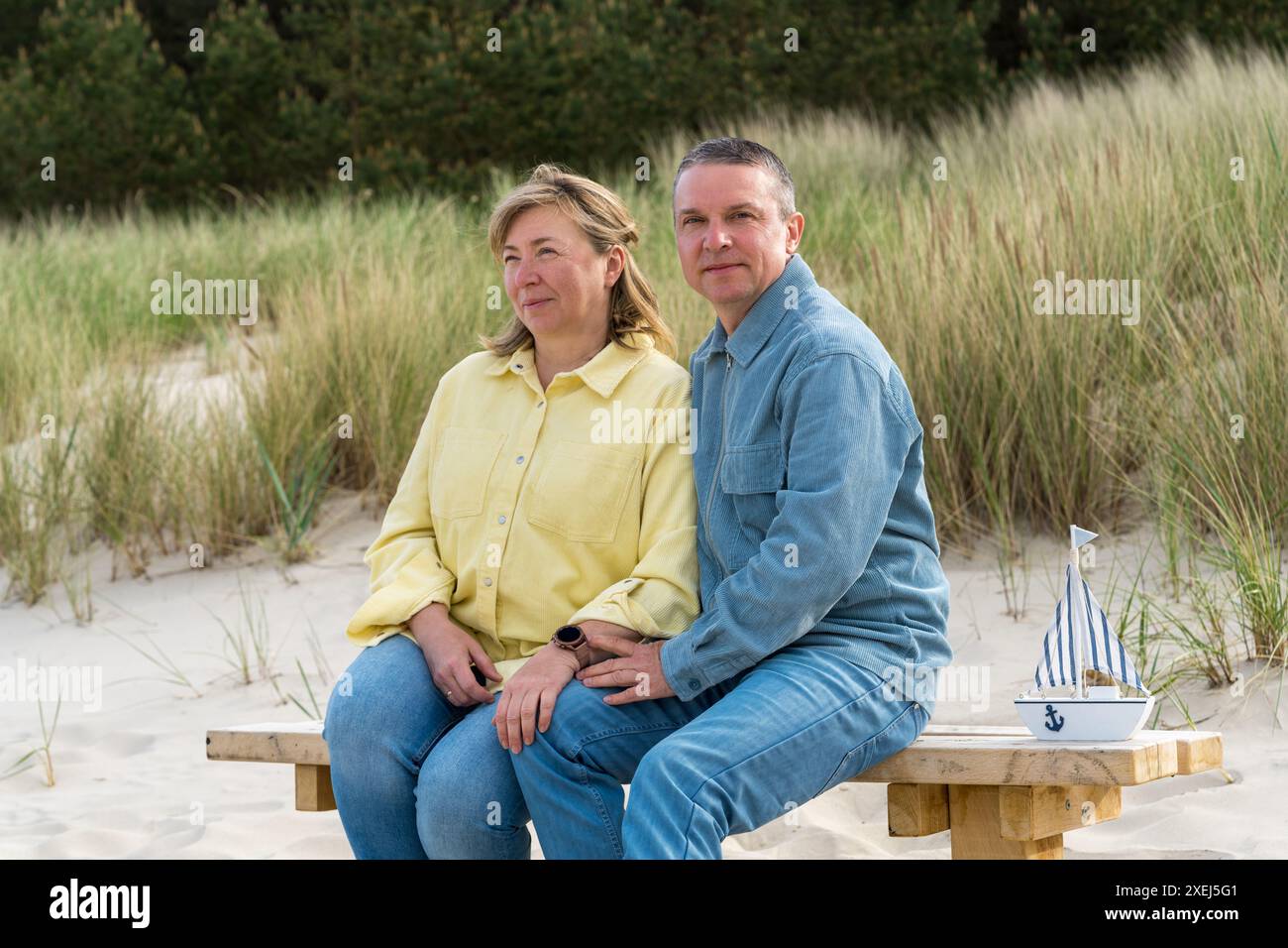 Ein Paar mittleren Alters im Urlaub auf dem Meer. Ein Mann und eine Frau genießen ihren Urlaub. Sie sitzen auf einer Bank an der Ostsee, mit Dünen in Backgr Stockfoto