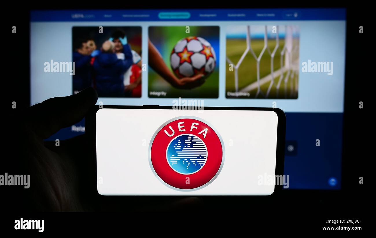 Person, die ein Mobiltelefon mit dem Logo der Union Europäischer Fußballverbände (UEFA) vor der Webseite hält. Schwerpunkt auf dem Display des Telefons. Stockfoto