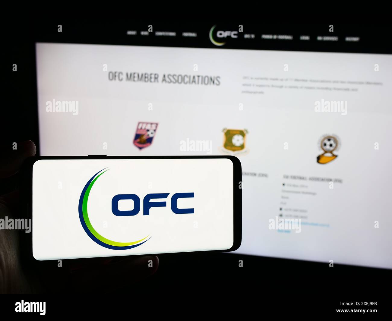 Person, die ein Mobiltelefon mit dem Logo des Fußballverbandes Oceania Football Confederation (OFC) vor der Webseite hält. Schwerpunkt auf dem Display des Telefons. Stockfoto