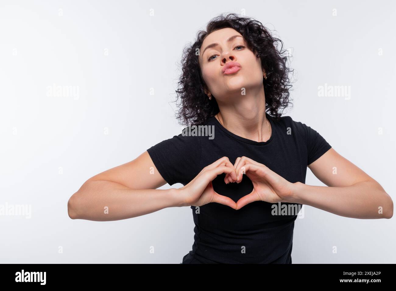 Erwachsene gelockte europäische Frau in schwarzem T-Shirt mit Herzgeste Stockfoto