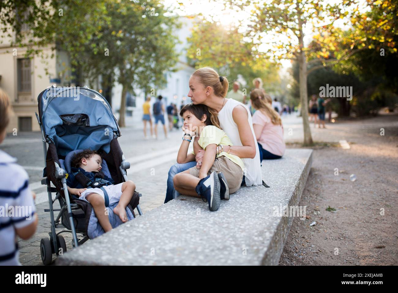 Eine Mutter und ihre beiden Söhne genießen einen gemütlichen Moment draußen in der Stadt, wobei ein Sohn in einem Kinderwagen schläft und der andere sich gegen seine Mutter lehnt. Stockfoto