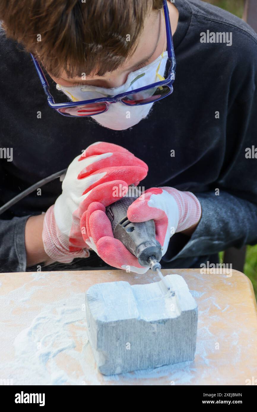 Junge, die Schutzkleidung tragen und mit einem Elektrowerkzeug einen Steinblock schnitzen Stockfoto