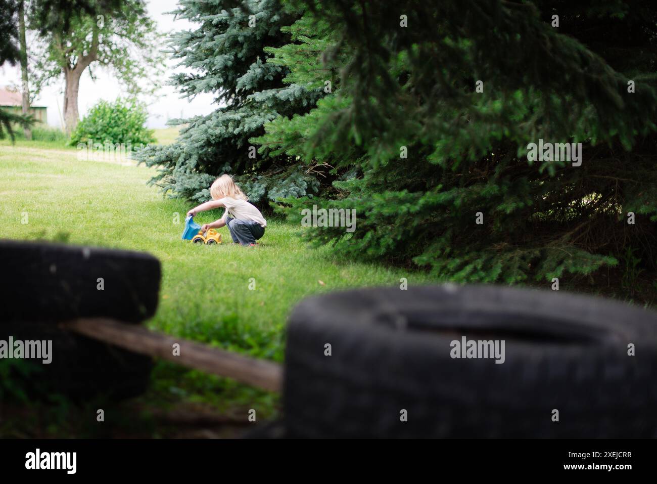 Kind, das mit einem Spielzeugwagen auf einem grasbewachsenen Rasen mit immergrünen Bäumen spielt Stockfoto