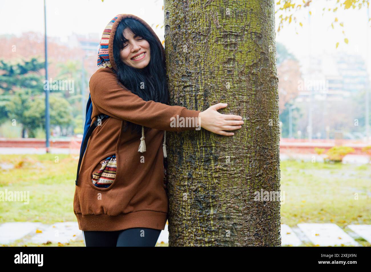 Frau, die glücklich ist, mit der Natur in der Stadt in Verbindung zu treten, sie umarmt Bäume und lächelt, Konzept der Rettung des Planeten Erde und der Pflege der natürlichen Ressourcen Stockfoto