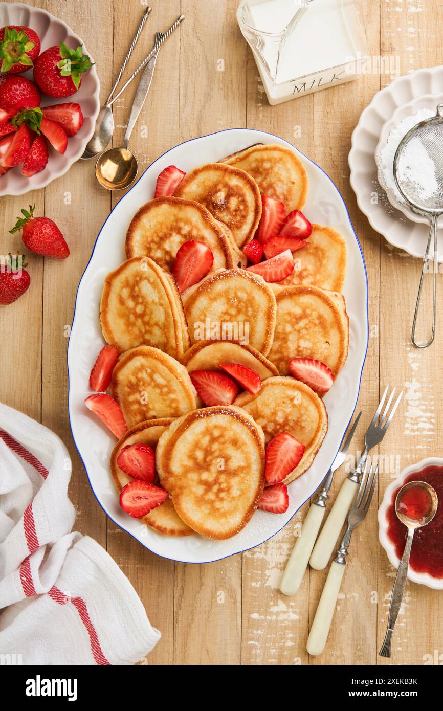 Pfannkuchen gestapelt auf weißer Keramikplatte mit Zuckerpulver, Marmelade und Erdbeere auf altem rustikalen hellen hölzernen Hintergrund. Idee, Frühstück und Mittagessen zu servieren Stockfoto