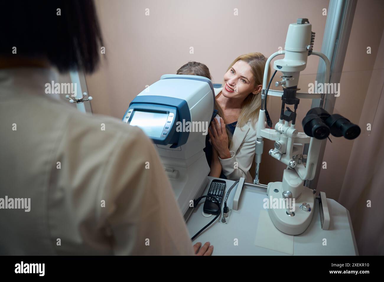 Eine Blondine mit einer kleinen Tochter sitzt an einem Autorefraktometer Stockfoto