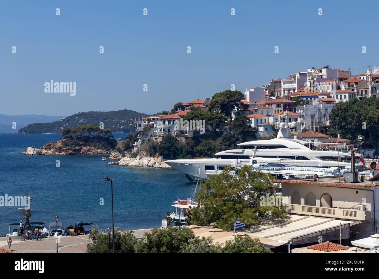 20. juni 2024, Skiathos, Griechenland Ein malerischer Yachthafen mit Booten, die an einer ruhigen Küstenstadt vor Anker liegen und sich auf sanften Hügeln unter klarem blauem Himmel befinden Stockfoto