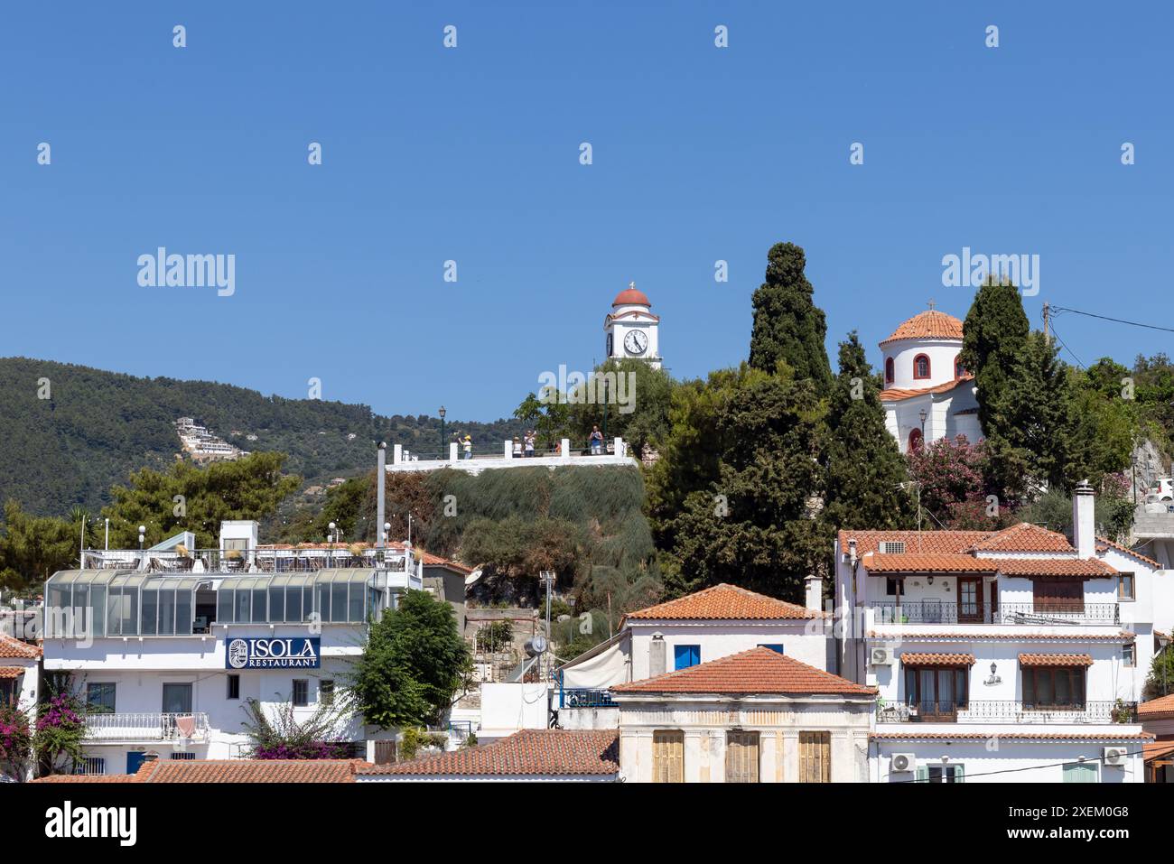 20. juni 2024, Skiathos, Griechenland Ein malerischer Yachthafen mit Booten, die an einer ruhigen Küstenstadt vor Anker liegen und sich auf sanften Hügeln unter klarem blauem Himmel befinden Stockfoto