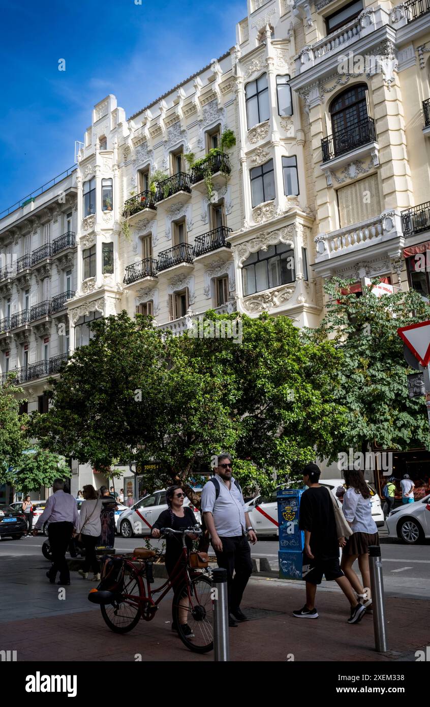 Straßenszene mit Fußgängern und Radfahrern, geparkten Fahrzeugen und einem kunstvollen Wohngebäude an einem sonnigen Tag in Madrid; Madrid, Spanien Stockfoto