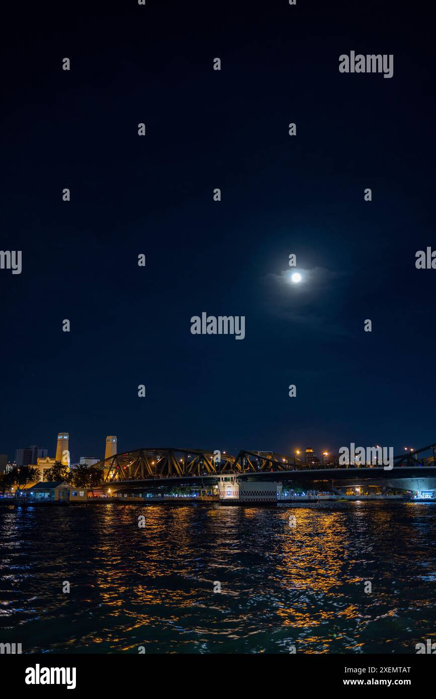 Nächtlicher Blick auf die Chao Phraya Sky Park Bridge über den Chao Phraya Fluss in Bangkok, während sich der Mond auf dem Wasser spiegelt; Bangkok, Thailand Stockfoto