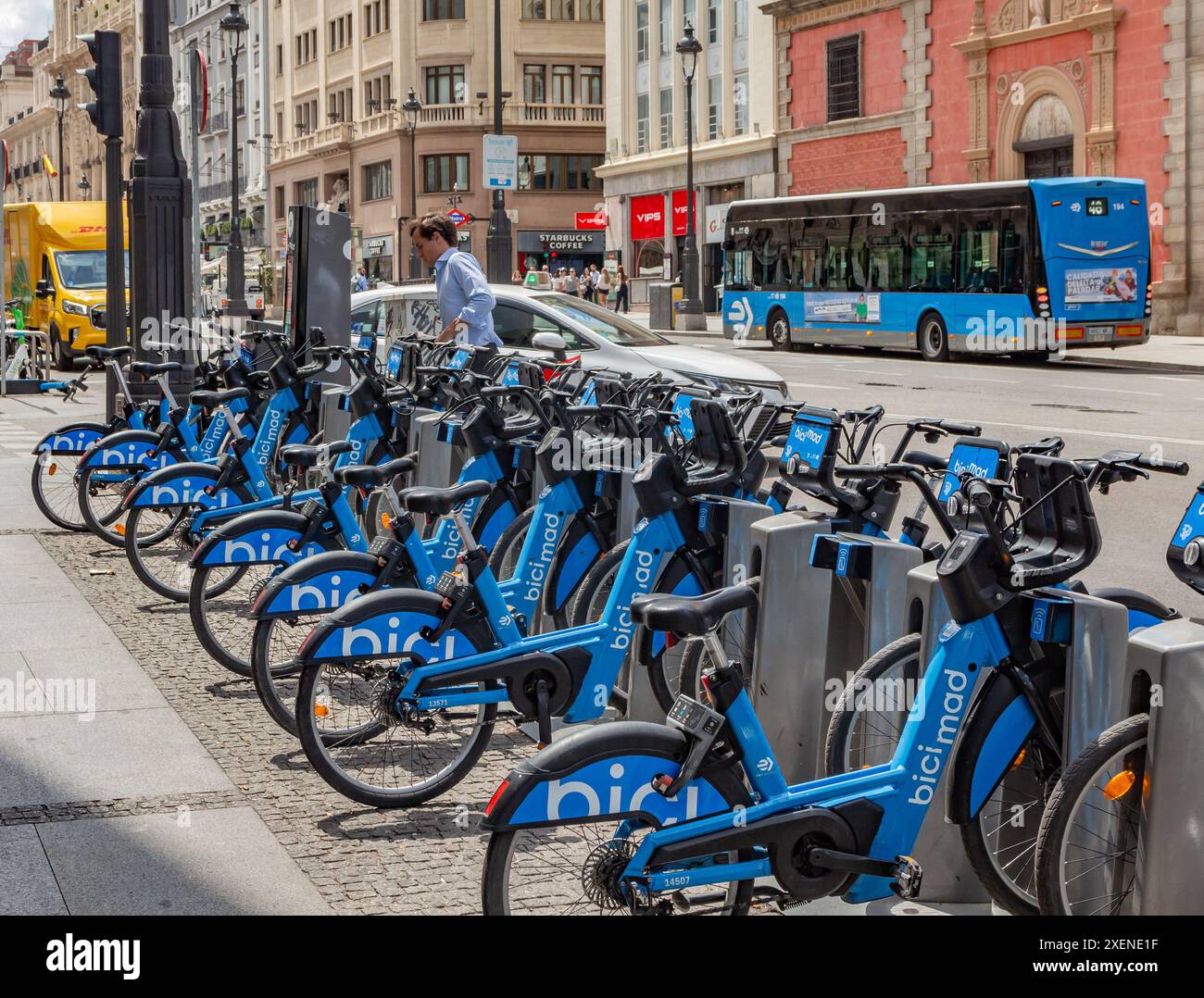 06.19.2024, Madrid, Spanien: Mann in blauem Hemd parkt ein BiciMAD-Elektrofahrrad an einer Dockingstation in einer Straße mit Bus und Verkehr Stockfoto