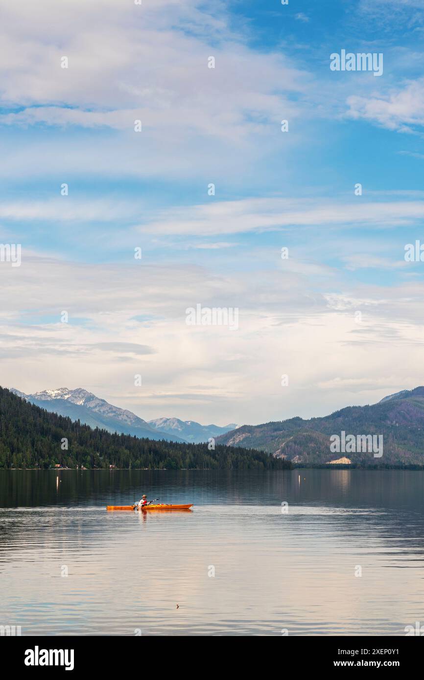 Ein vertikales Bild eines Mannes in einem orangen Kajak auf dem Lake Wenatchee im Bundesstaat Washington mit Kaskadenzügen in der Ferne. Stockfoto