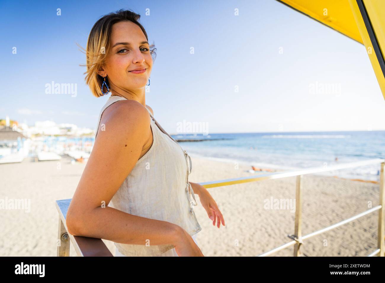 Eine Frau steht am Strand mit einem gelben Schirm hinter sich. Sie lächelt und posiert für ein Foto Stockfoto