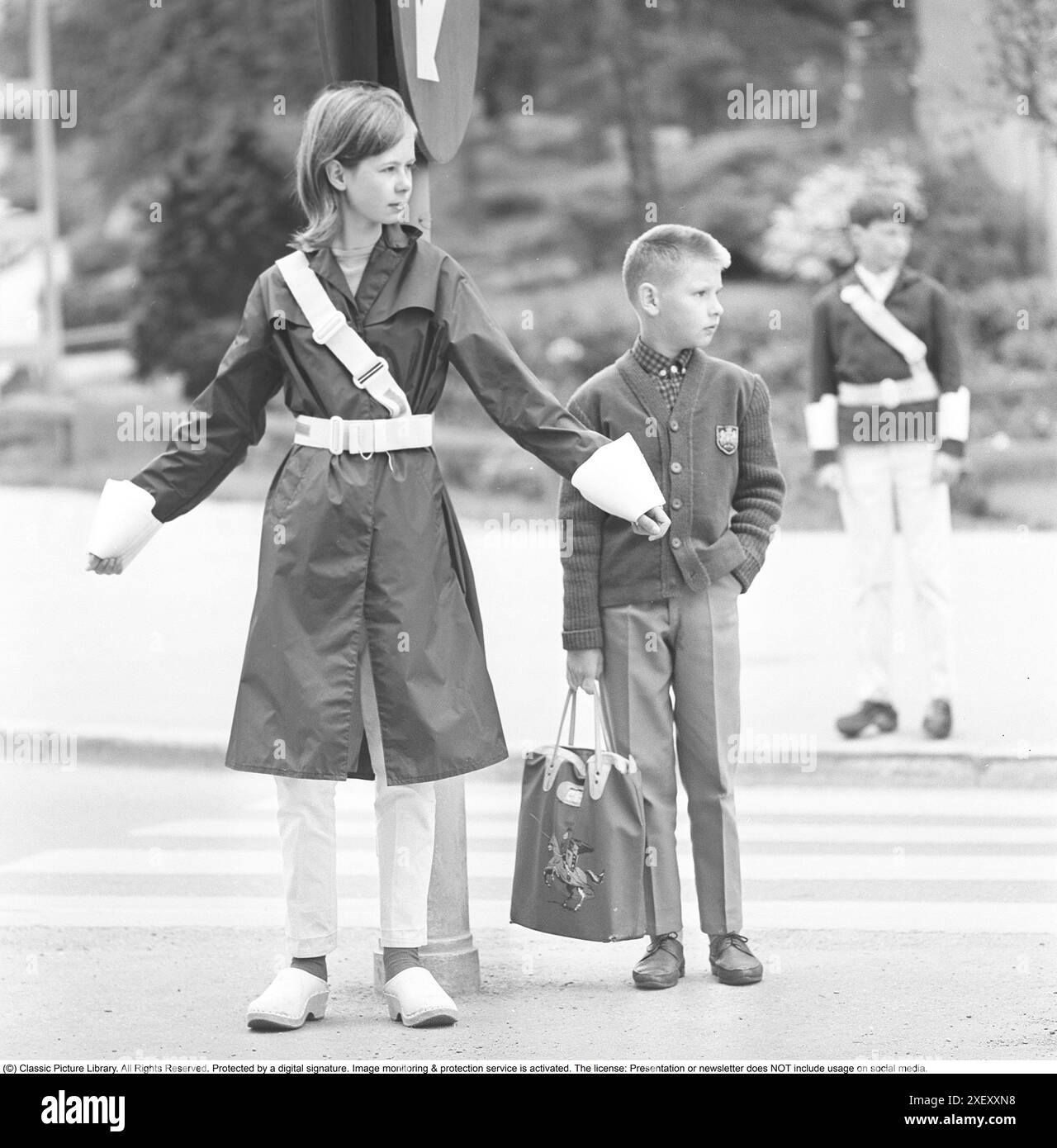 Junior-Sicherheitspatrouille in den 1960er Jahren Ein Mädchen im Teenageralter steht mit ausgestreckten Armen und signalisiert den Fußgängern, an der Kreuzung anzuhalten. Wenn die Autos vorbei sind oder angehalten haben, können die Kinder über die Straße laufen. Die Teilnahme an der Junior-Sicherheitspatrouille war freiwillig für die Schülerinnen und Schüler und die Belohnung bestand manchmal darin, auf kürzeren Touren und Exkursionen zu gehen. Heute ist der Betrieb in vielen Gemeinden geschlossen. Schweden 1964 Stockfoto