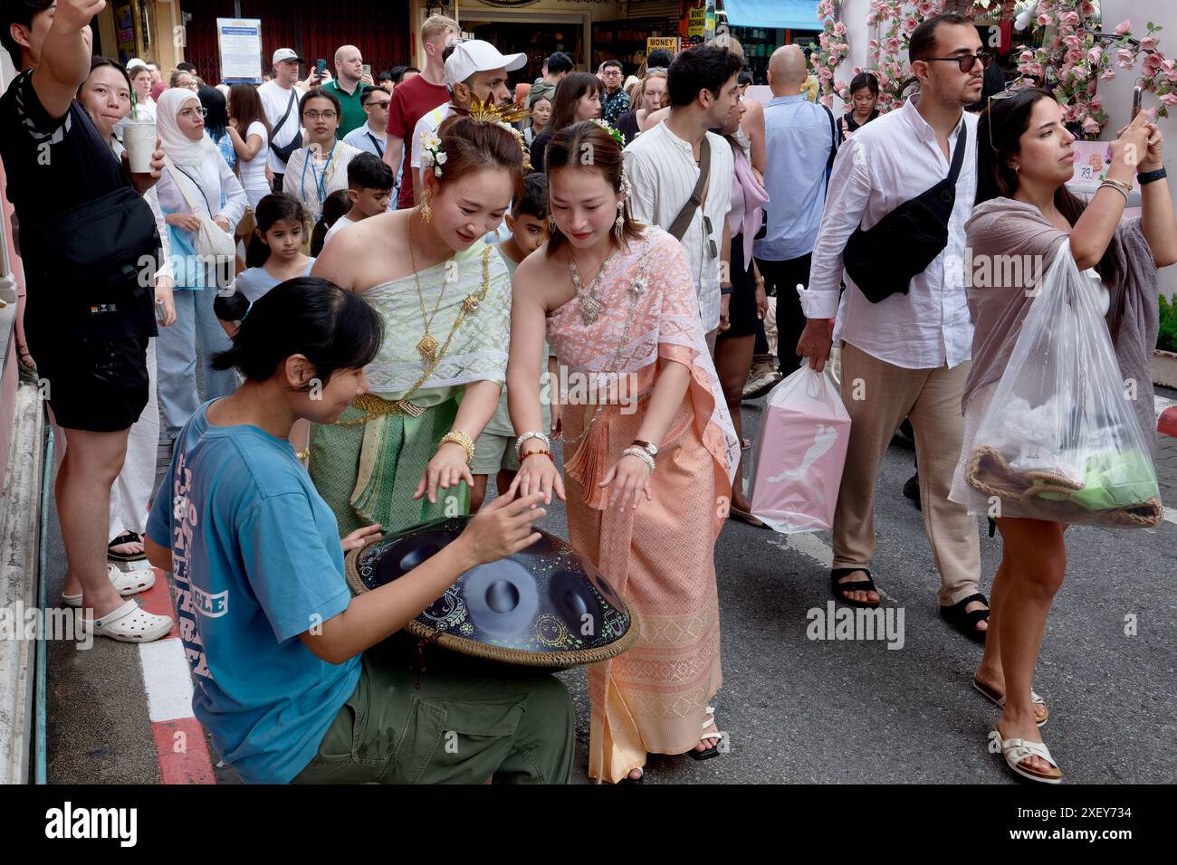 Thailändische Touristen in der Altstadt von Phuket in traditioneller thailändischer Tracht und sprechen mit einem Straßenmusiker, der eine Handpfanne spielt, einer Handtrommel aus Metall Stockfoto